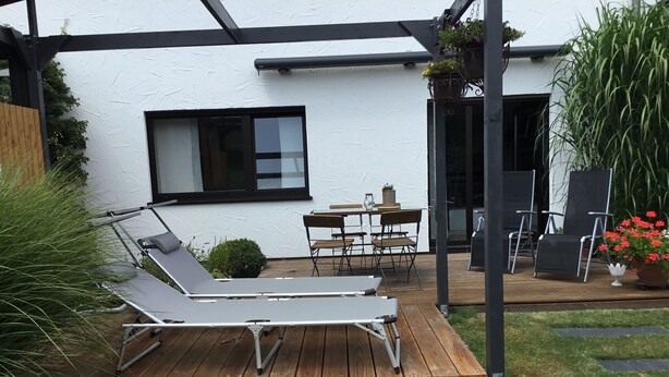 Une terrasse en bois avec un fauteuil de relaxation, une table et des chaises. On aperçoit une fenêtre et une porte à l’arrière-plan.
