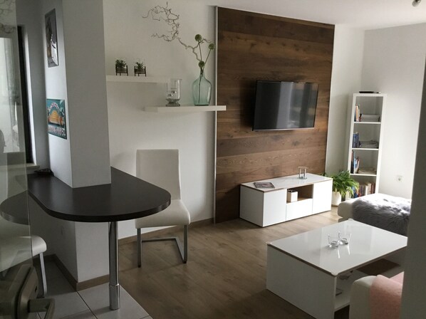 A modern living room with a dark wooden accent wall, a flat-screen TV mounted above a white console, and a white sofa. - Modern, sunny vacation apartment - quiet location, with large terrace (Altrich)