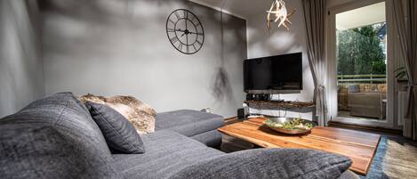 A modern living room with a grey sofa, a wooden coffee table, a flat-screen TV, and a large window with curtains.