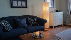 A living room with a dark blue sofa, patterned cushions, a wooden coffee table with a candle, a tall floor lamp, a white dresser, and framed artwork on the wall.