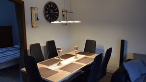 A dining area with a rectangular table, six chairs, a wall clock, and a bed in the background.