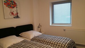 Une chambre avec deux lits, un radiateur, une fenêtre avec des stores et une lampe fixée au mur.