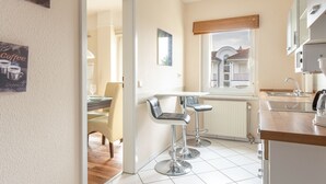 A modern kitchen with two bar stools, a dining area with a table and chairs, and a view of a building outside. - 3-room apartment Haus Godewind 10 in Binz, balcony, parking lot, beach chair on the beach (Binz)