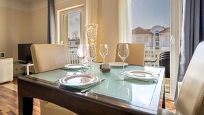 A dining table set with plates, cutlery, and wine glasses, with a view of buildings through large windows. - 3-room apartment Haus Godewind 10 in Binz, balcony, parking lot, beach chair on the beach (Binz)