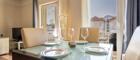 Mesa de comedor dispuesta con platos, cubiertos y copas de vino, con vistas a edificios a través de amplias ventanas.
