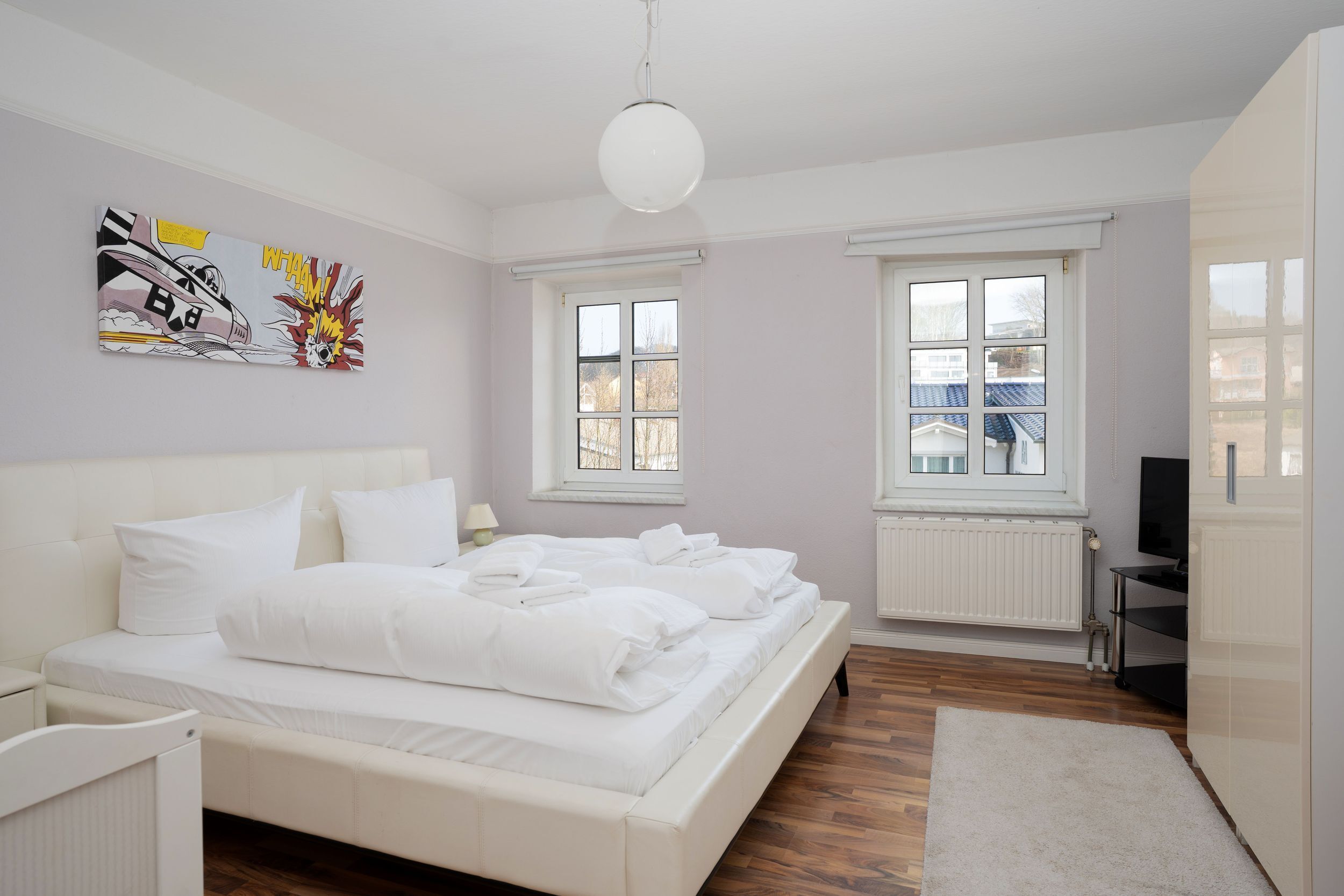 Ein modernes Schlafzimmer mit einem großen Bett, zwei Fenstern, einer Heizung und einem Fernseher.