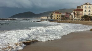 Uma praia com ondas quebrando na costa, edifícios ao fundo e um céu nublado.