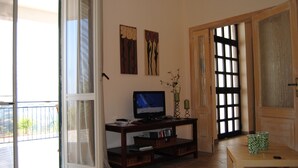Uma sala de estar com televisão sobre um suporte de madeira, uma porta de vidro deslizante que dá para uma varanda e uma pequena mesa com duas chávenas verdes.