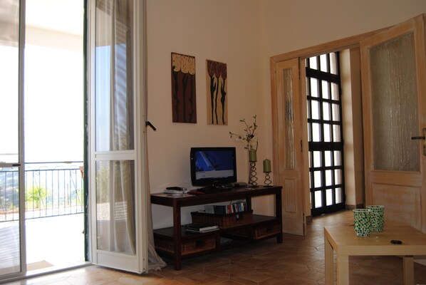 A living room with a television on a wooden stand, a sliding glass door leading to a balcony, and a small table with two green cups.