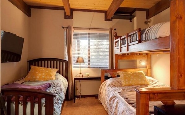 A room with two wooden bunk beds, a desk with a lamp, and a television on the wall.