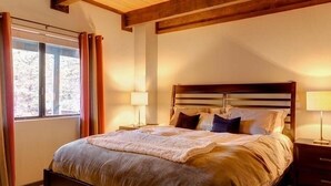 A bedroom with a large bed, wooden ceiling beams, a window with curtains, and bedside lamps.