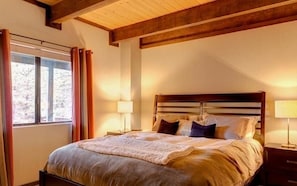 Quarto com cama grande, vigas de madeira no teto, janela com cortinas e abajures.