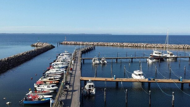 A marina with boats docked along a wooden pier, a rocky breakwater, and calm water.