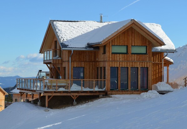 A wooden chalet with a balcony, large windows, and a snow-covered roof.
