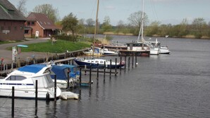 A marina with boats docked at wooden piers, a brick building with a green roof, and a grassy area with trees in the background. - At the ice cream parlor, Germany (Pahlen)