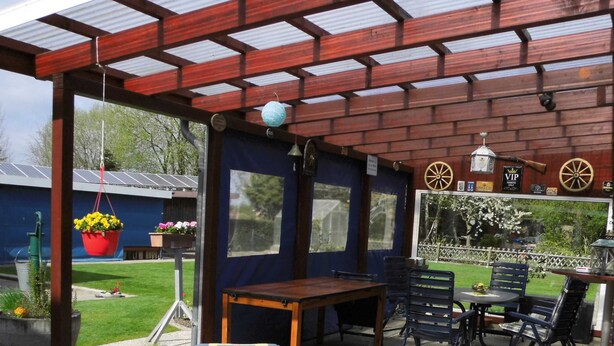 A covered patio area with a table and chairs, a bench, and hanging flower baskets. - At the ice cream parlor, Germany (Pahlen)