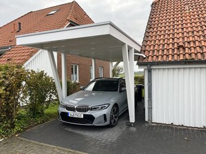 Een zilverkleurige BMW staat geparkeerd onder een witte carport in een woonwijk met rode dakpannen.