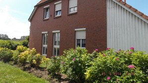 Ein zweistöckiges Backsteinhäuschen mit geneigtem Dach, weißen Fensterläden und einem Garten mit blühenden Pflanzen.