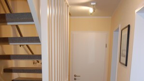 Ein schmaler Flur mit Holzdielen, einer weißen Tür und einer Treppe mit weißer Geländer.