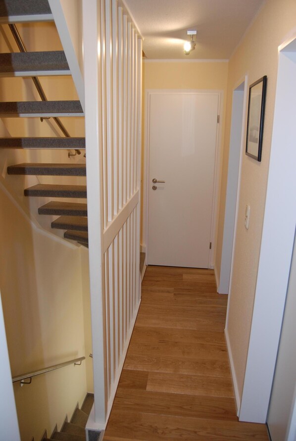 Ein schmaler Flur mit Holzdielen, einer weißen Tür und einer Treppe mit weißer Geländer.