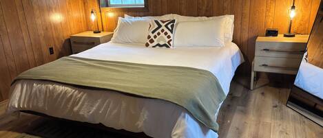 A wooden-paneled bedroom with a bed, two bedside tables, a nightstand, a window with blinds, and a wall-mounted light fixture.