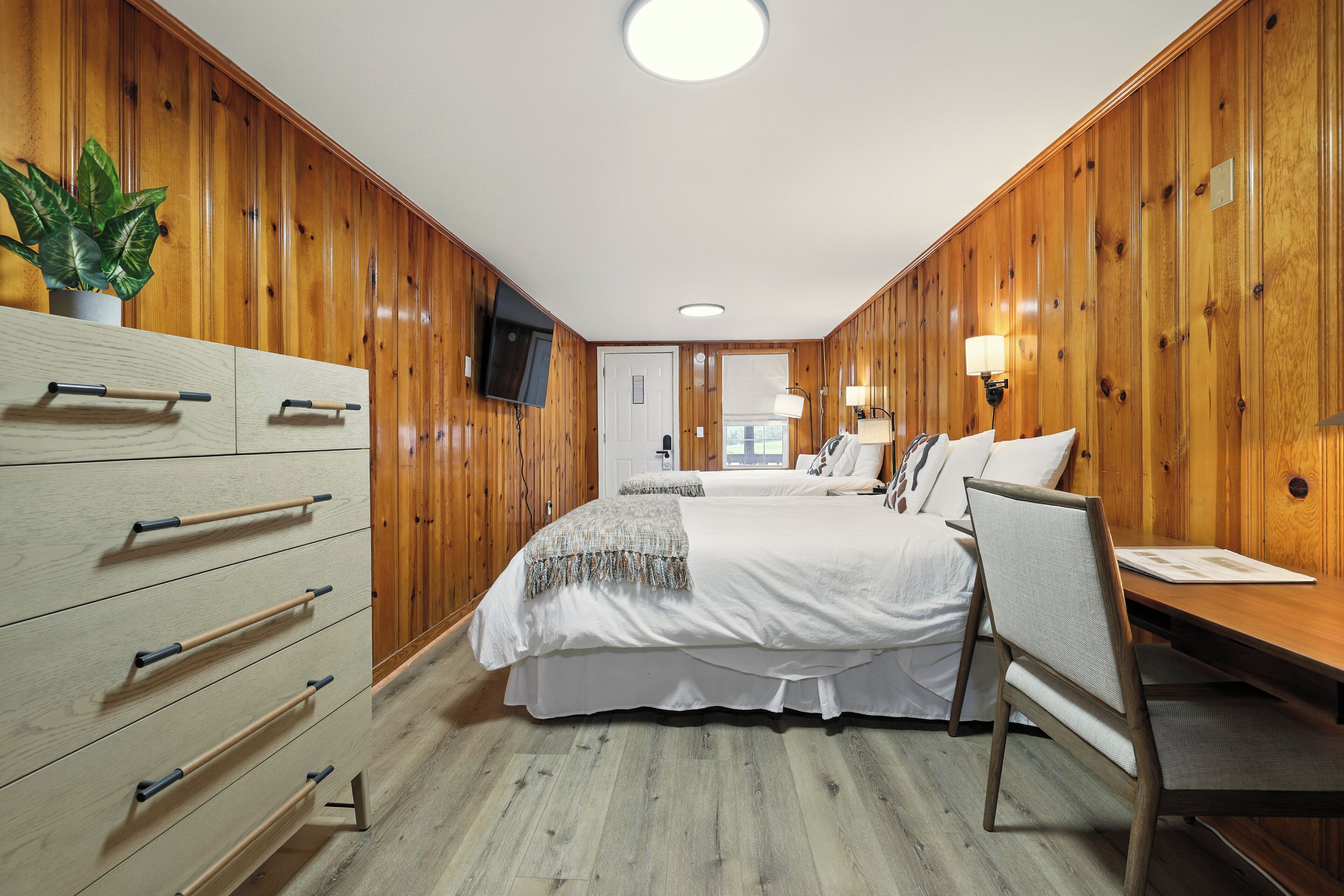 A bedroom with wooden paneling, a bed, a desk, a chair, a TV, and a plant.