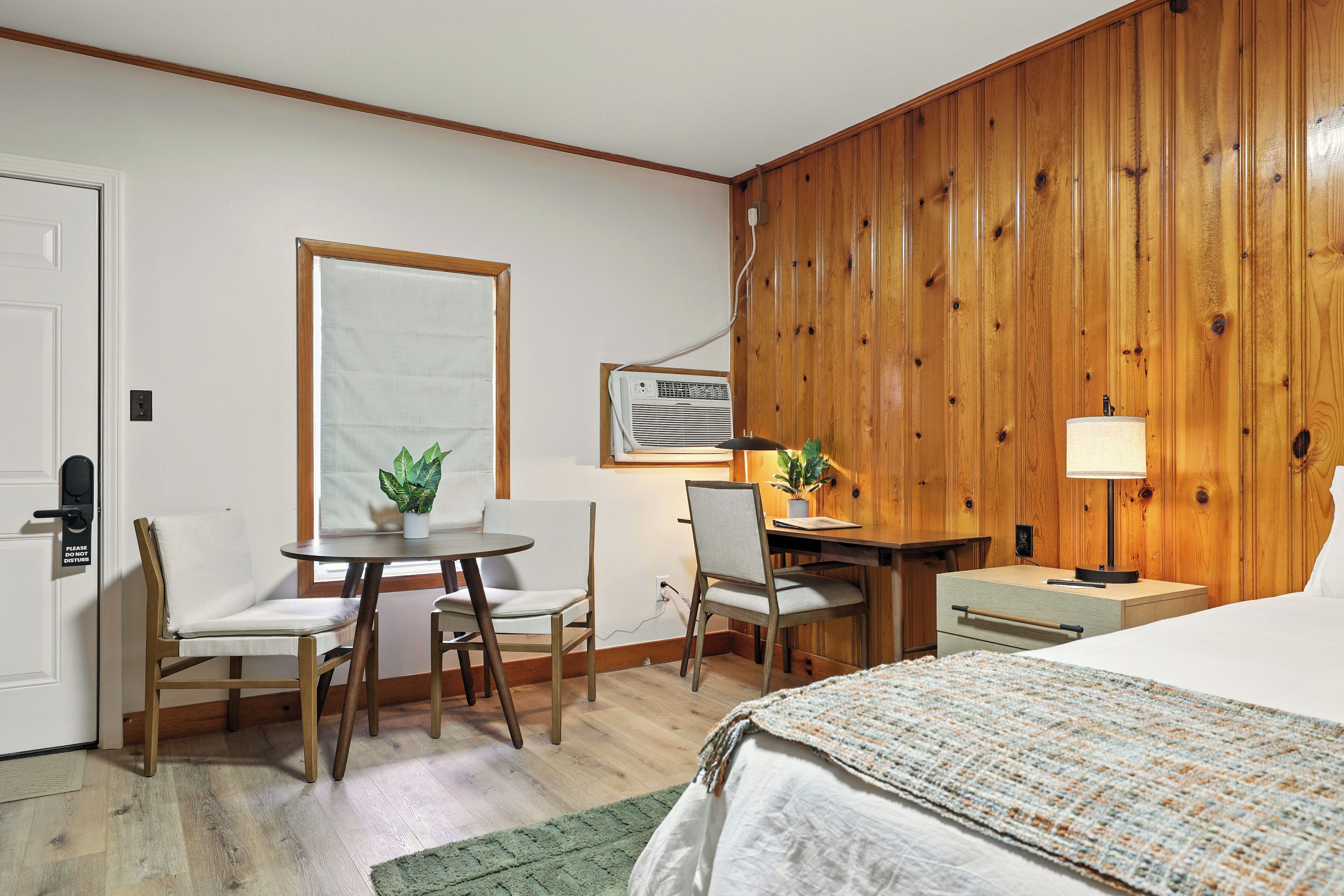 A hotel room with a bed, a desk, two chairs, a small table, a lamp, and a potted plant.