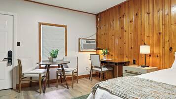 A hotel room with a bed, a desk, two chairs, a small table, a lamp, and a potted plant.