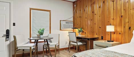A hotel room with a bed, a desk, two chairs, a small table, a lamp, and a potted plant.