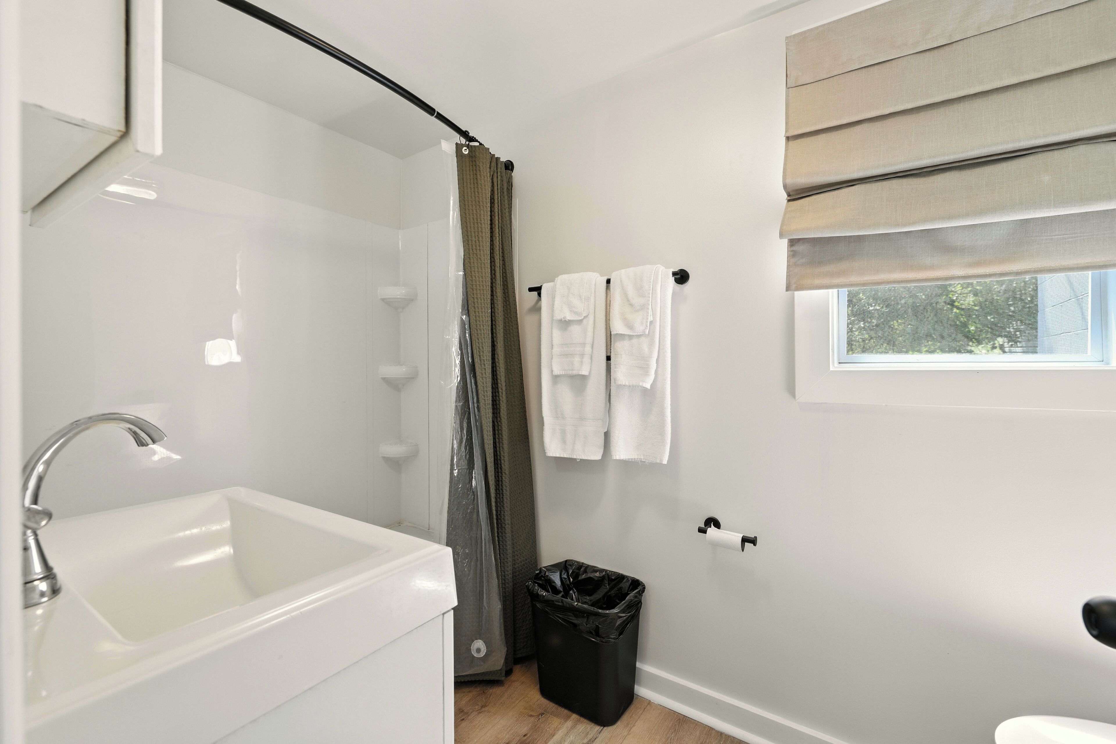 A bathroom with a white sink, a shower curtain, a black trash bin, and a window with a view of trees.