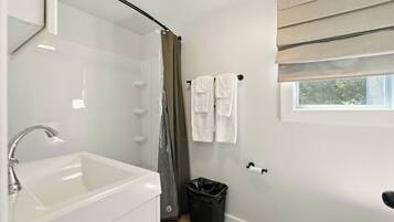 Un baño con un lavamanos blanco, cortina de ducha, un bote de basura negro y una ventana con vistas a árboles.