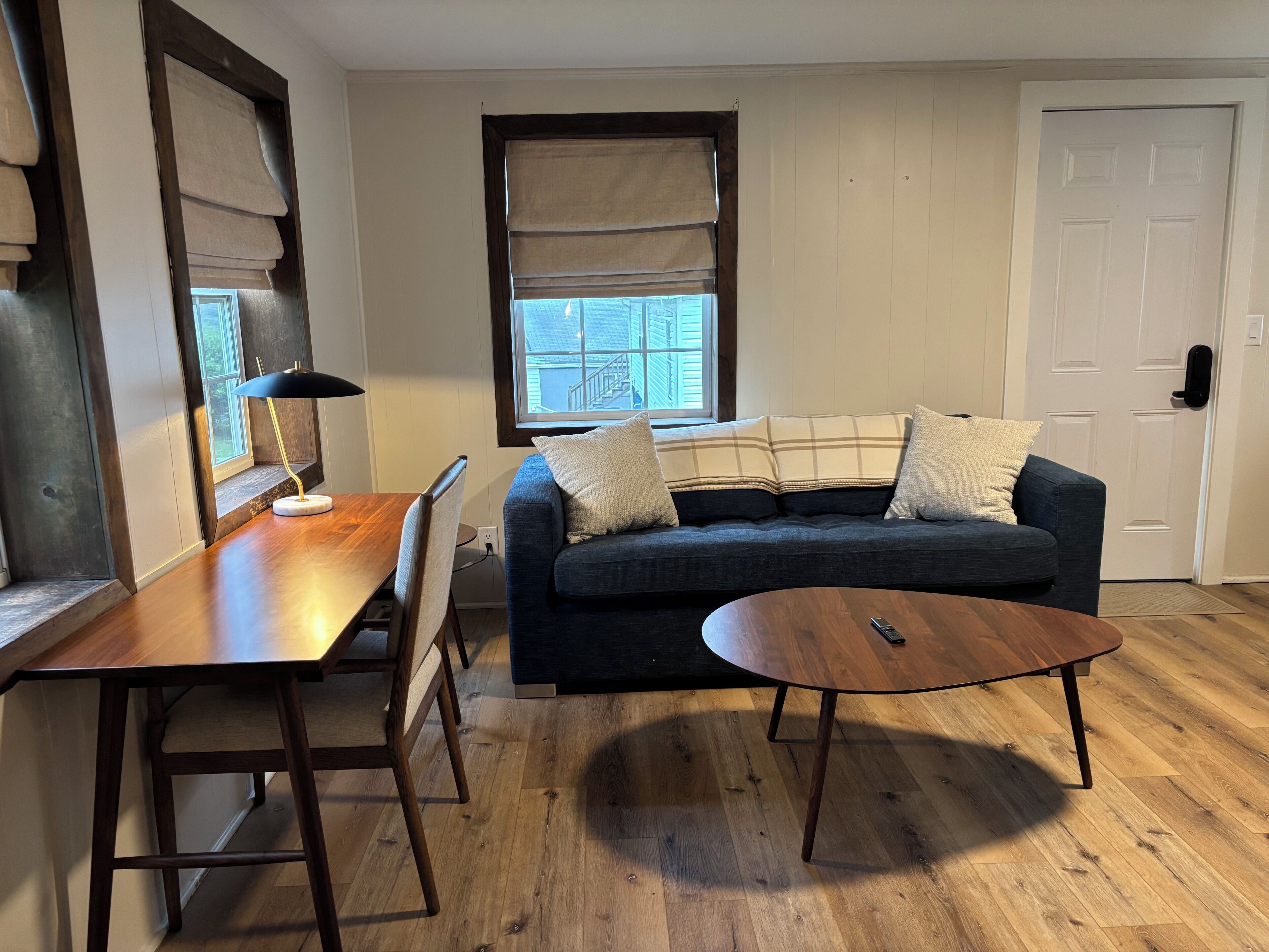 A living room with a wooden dining table, a dark blue sofa with pillows, a round coffee table, and a window with blinds.