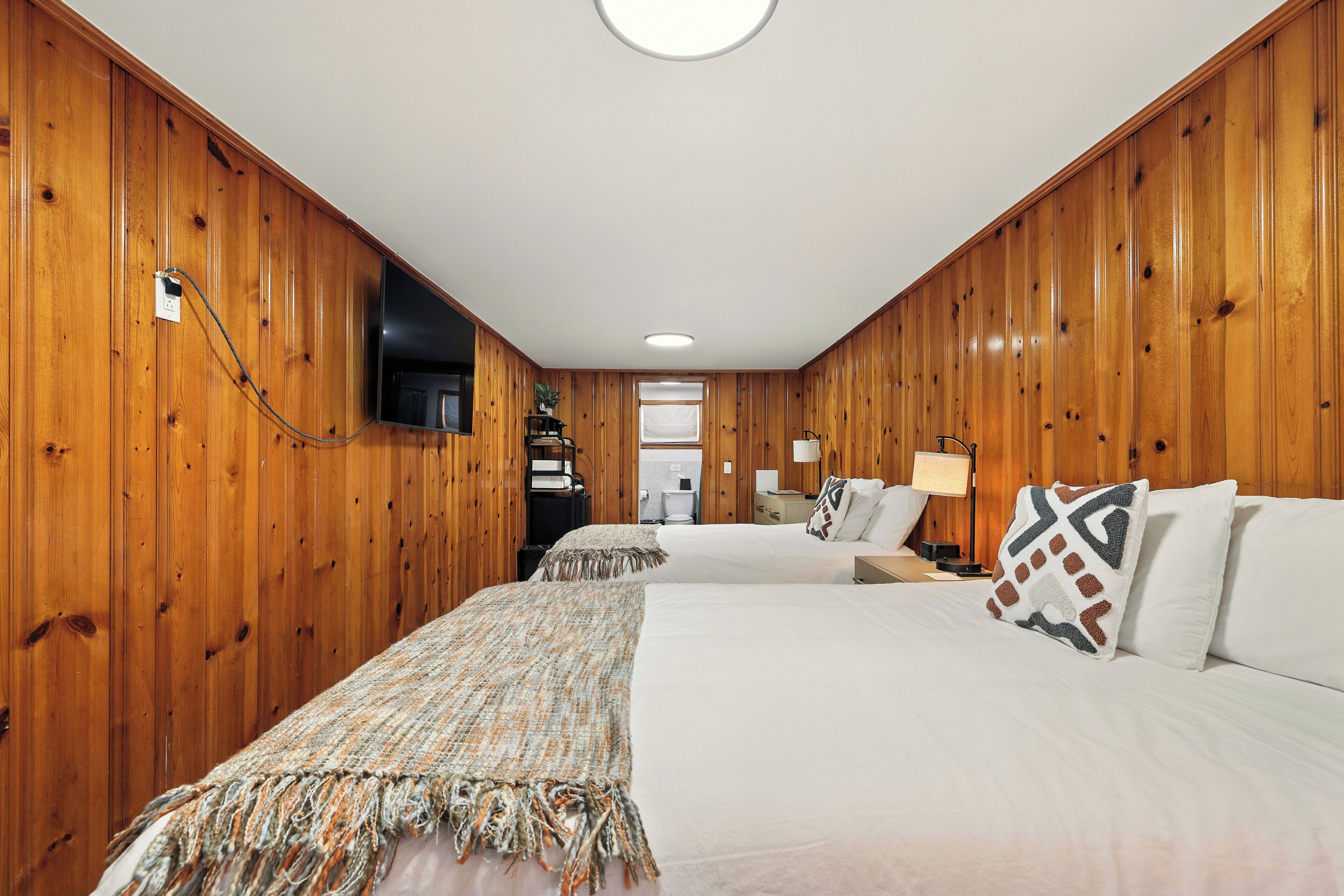 A hotel room with wooden paneling, two beds, a television, a desk with a lamp, and a window.