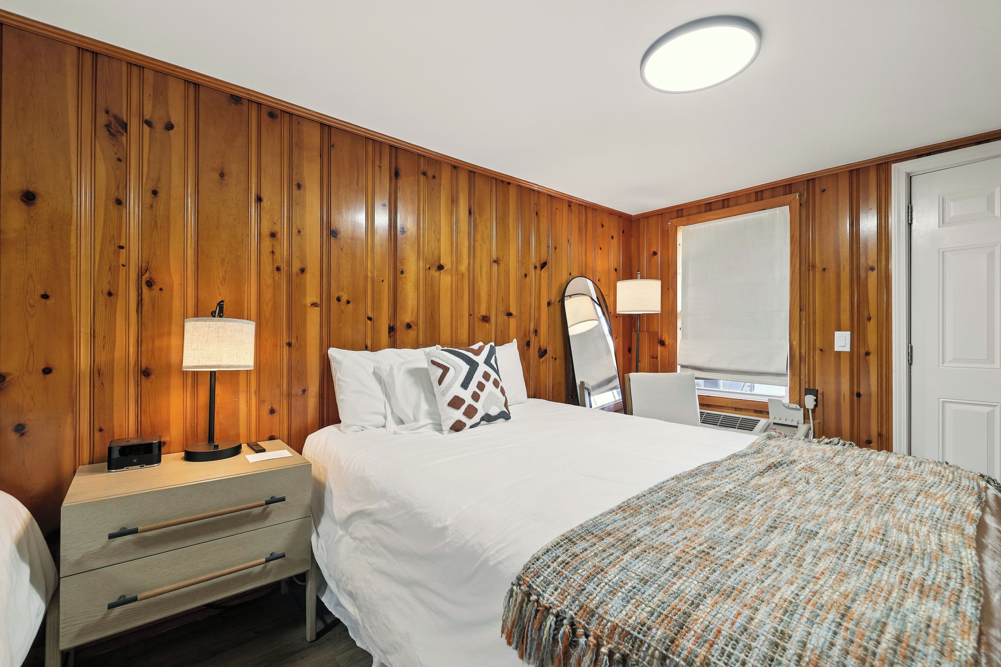A hotel room with wooden panel walls, a bed with white linens, a bedside table with a lamp, and a large mirror on the wall.