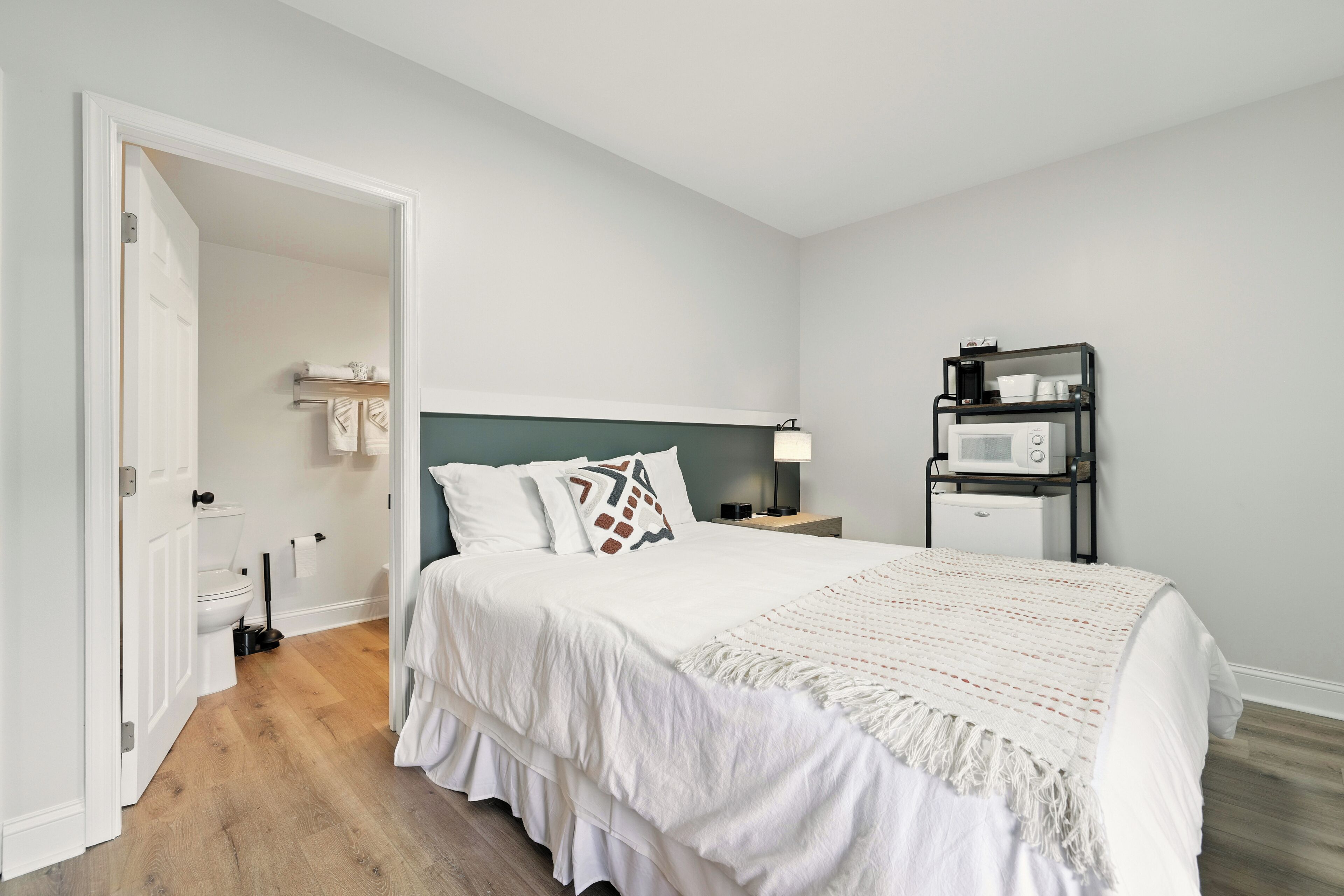 A bedroom with a bed, a nightstand, a lamp, a microwave, and a bathroom visible through an open door.