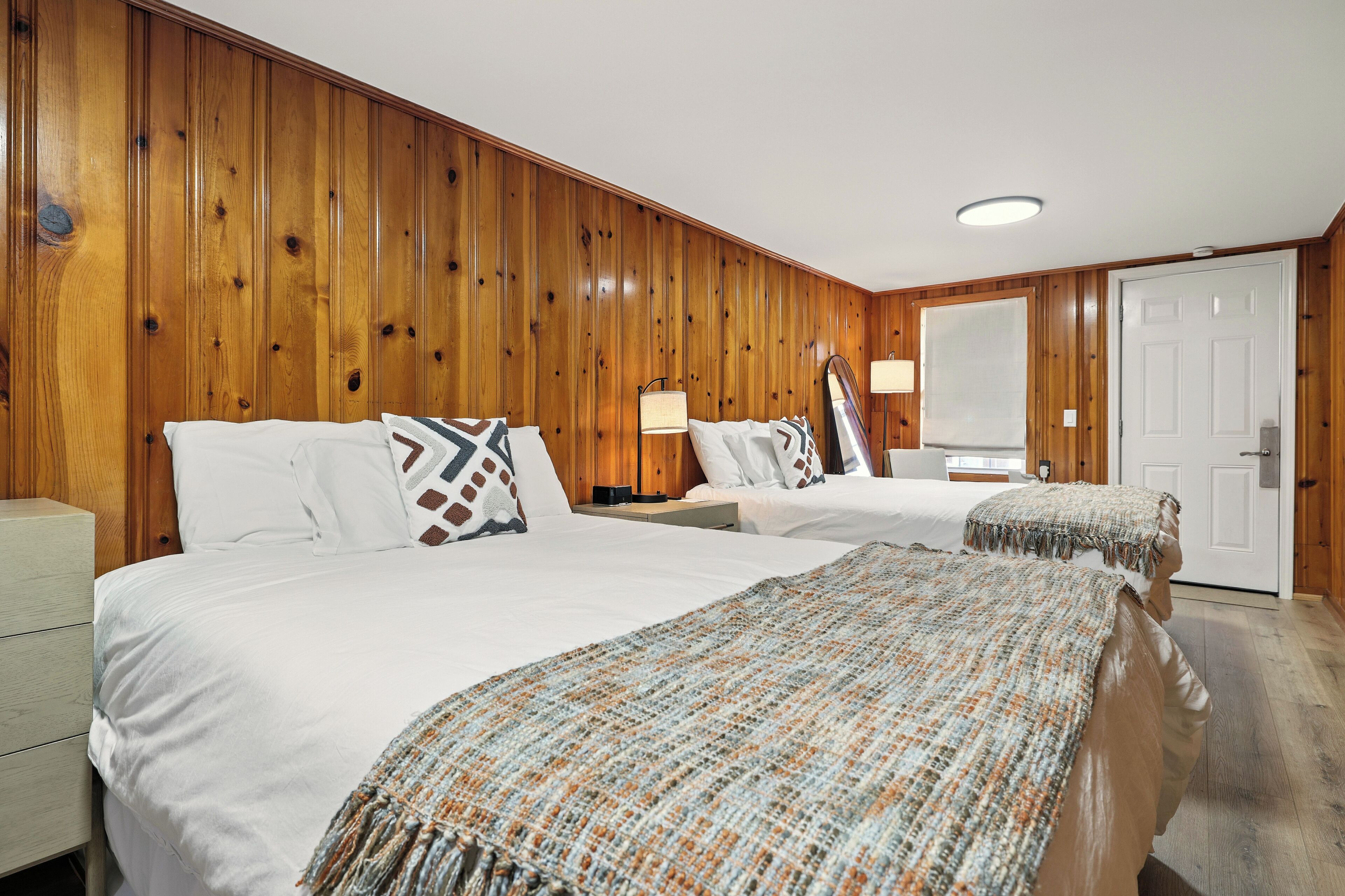 A hotel room with two beds, wooden paneling on the walls, a white door, and a nightstand with a lamp.