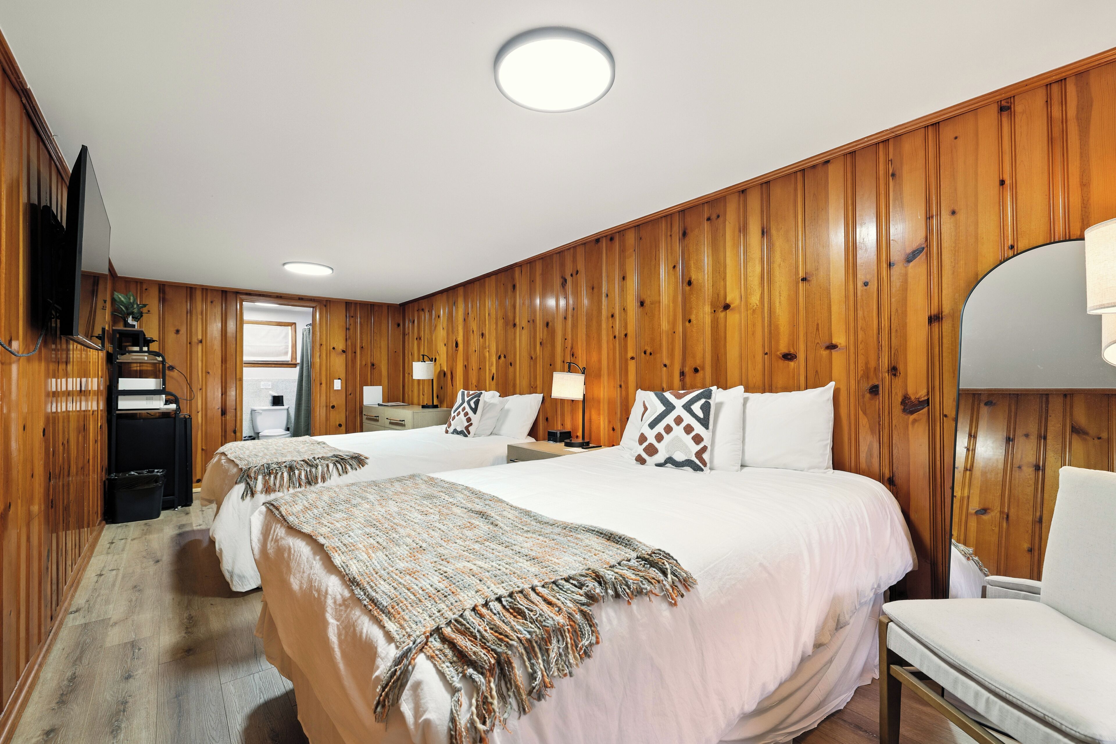 Quarto de hotel com painéis de madeira, duas camas com roupa branca, uma televisão, uma escrivaninha com abajur e um espelho.