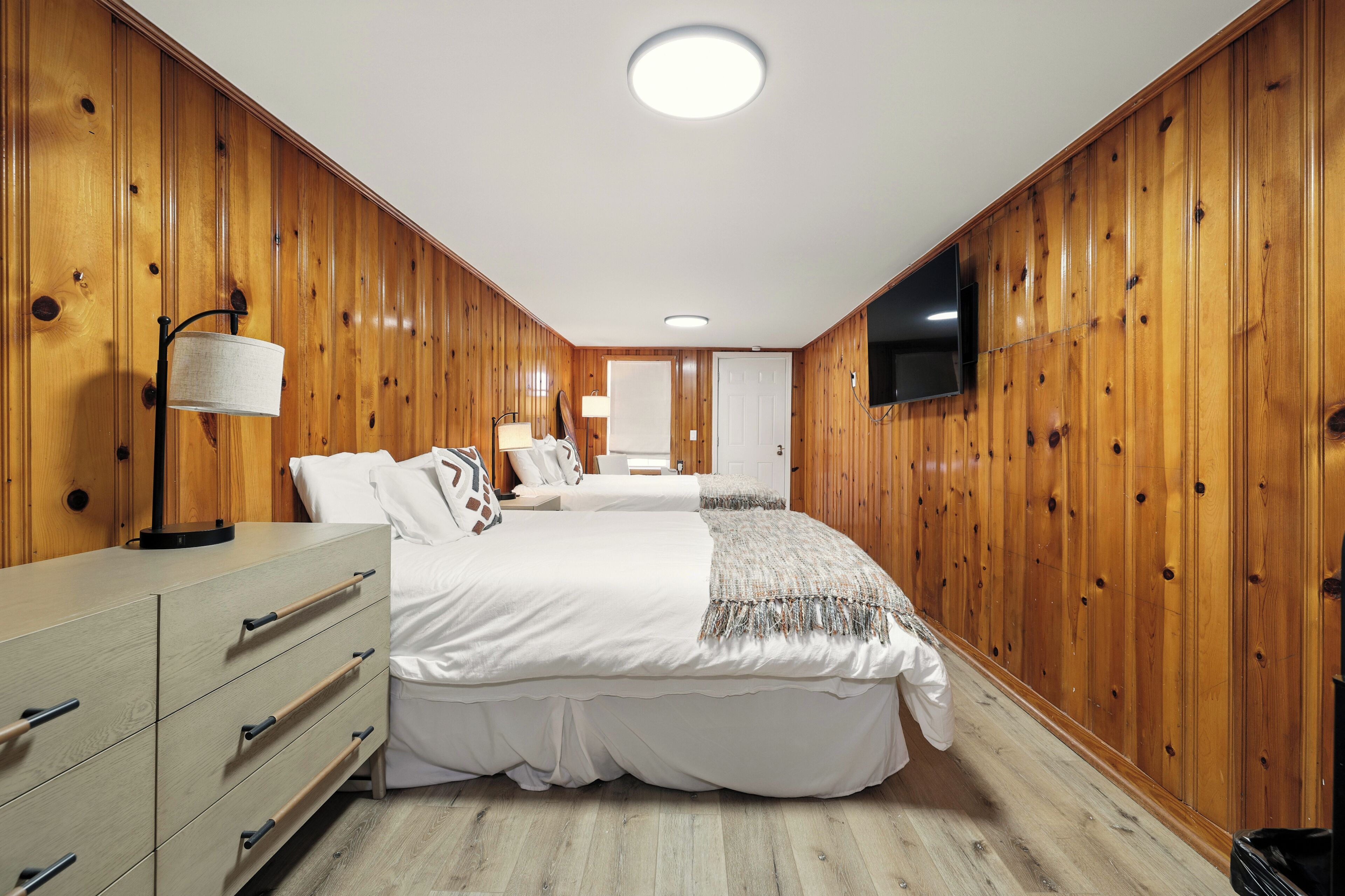 A bedroom with wooden paneling, a TV mounted on the wall, a bed with white bedding, and a bedside table with a lamp.