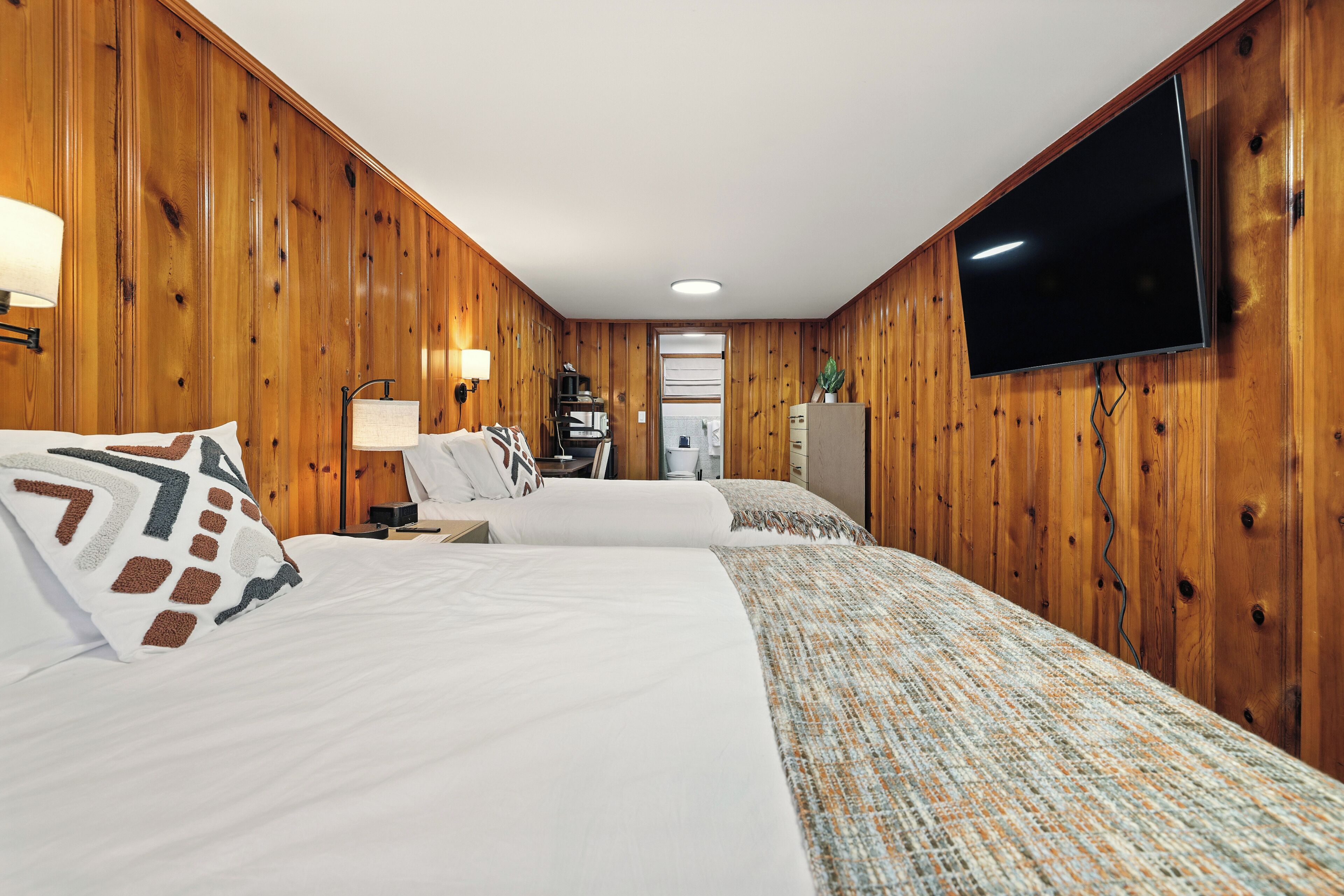 A hotel room with wooden panel walls, two beds with white linens, a flat-screen TV mounted on the wall, and a small desk with a lamp.