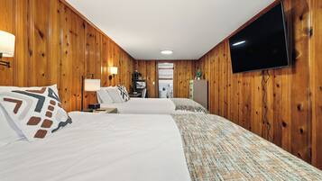 A hotel room with wooden panel walls, two beds with white linens, a flat-screen TV mounted on the wall, and a small desk with a lamp.