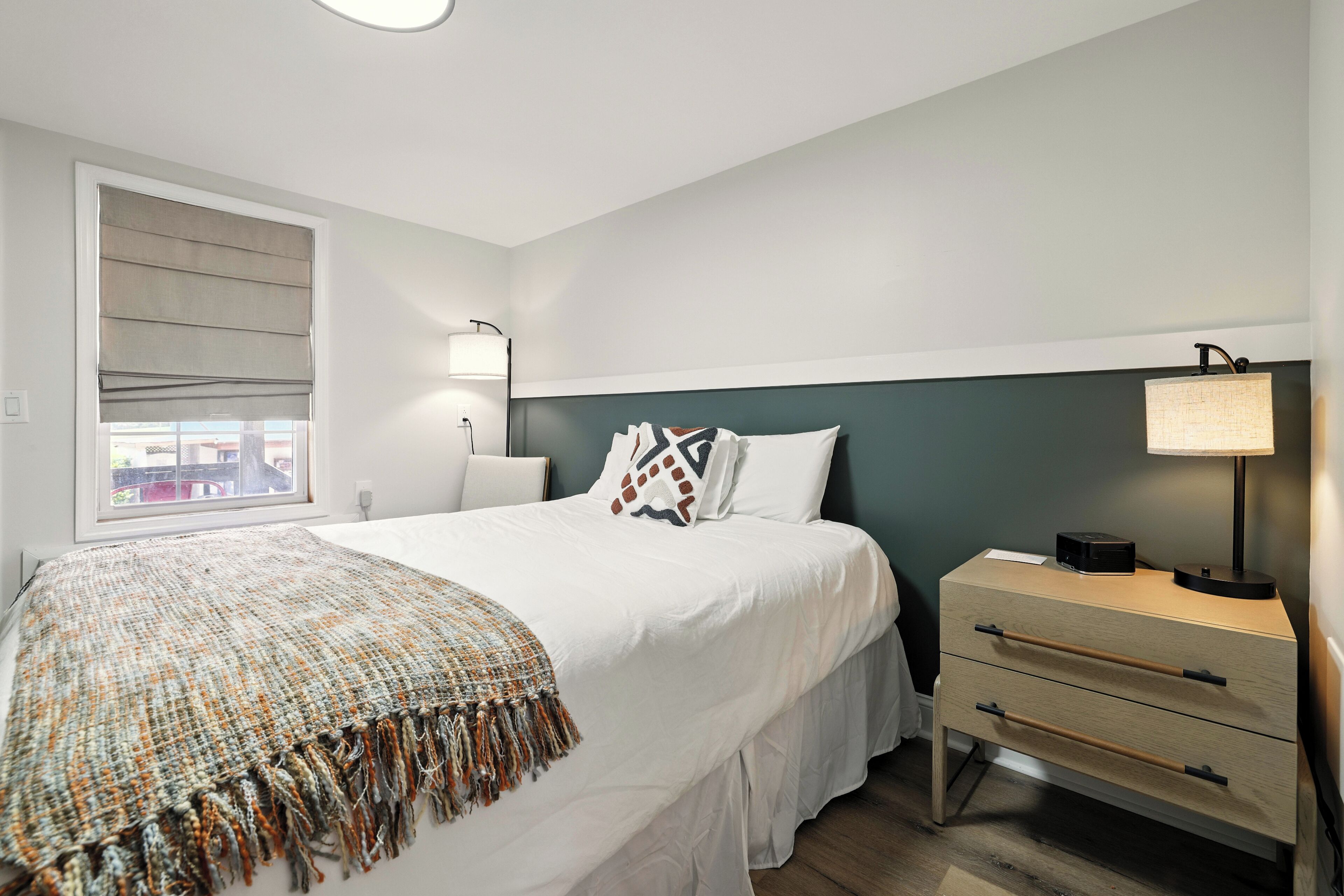 A bedroom with a bed, bedside table, lamp, and window with blinds.