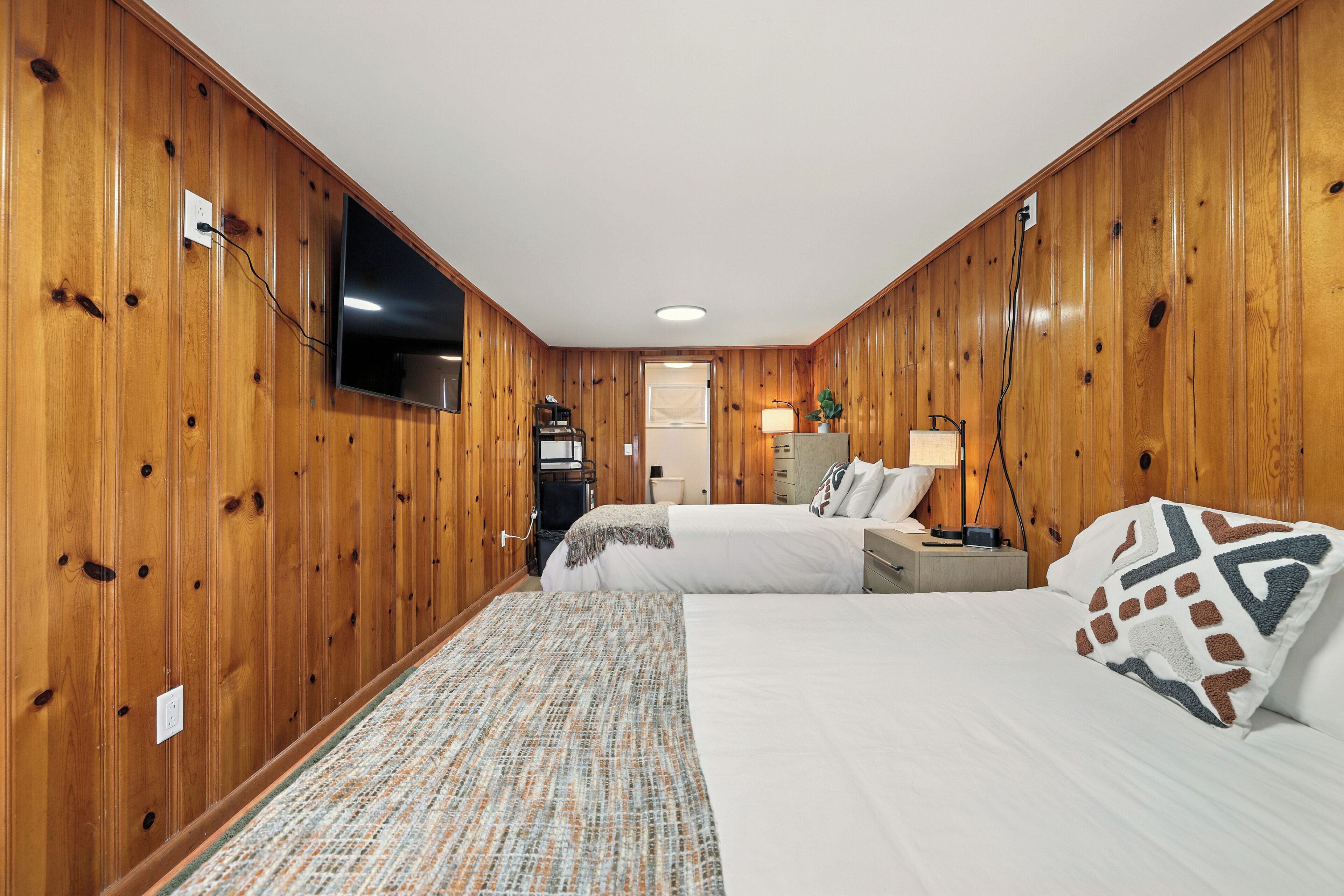A wooden-paneled room with two beds, a TV mounted on the wall, and a small desk with a lamp.