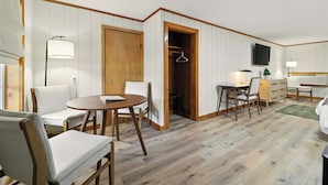 A room with a wooden floor, white walls, a wooden door, a table with chairs, a desk with a chair, a TV mounted on the wall, and a coat hanger in the closet. - Hillside (Luray)