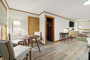 Una habitación con piso de madera, paredes blancas, una puerta de madera, una mesa con sillas, un escritorio con una silla, un televisor montado en la pared y un perchero en el armario.