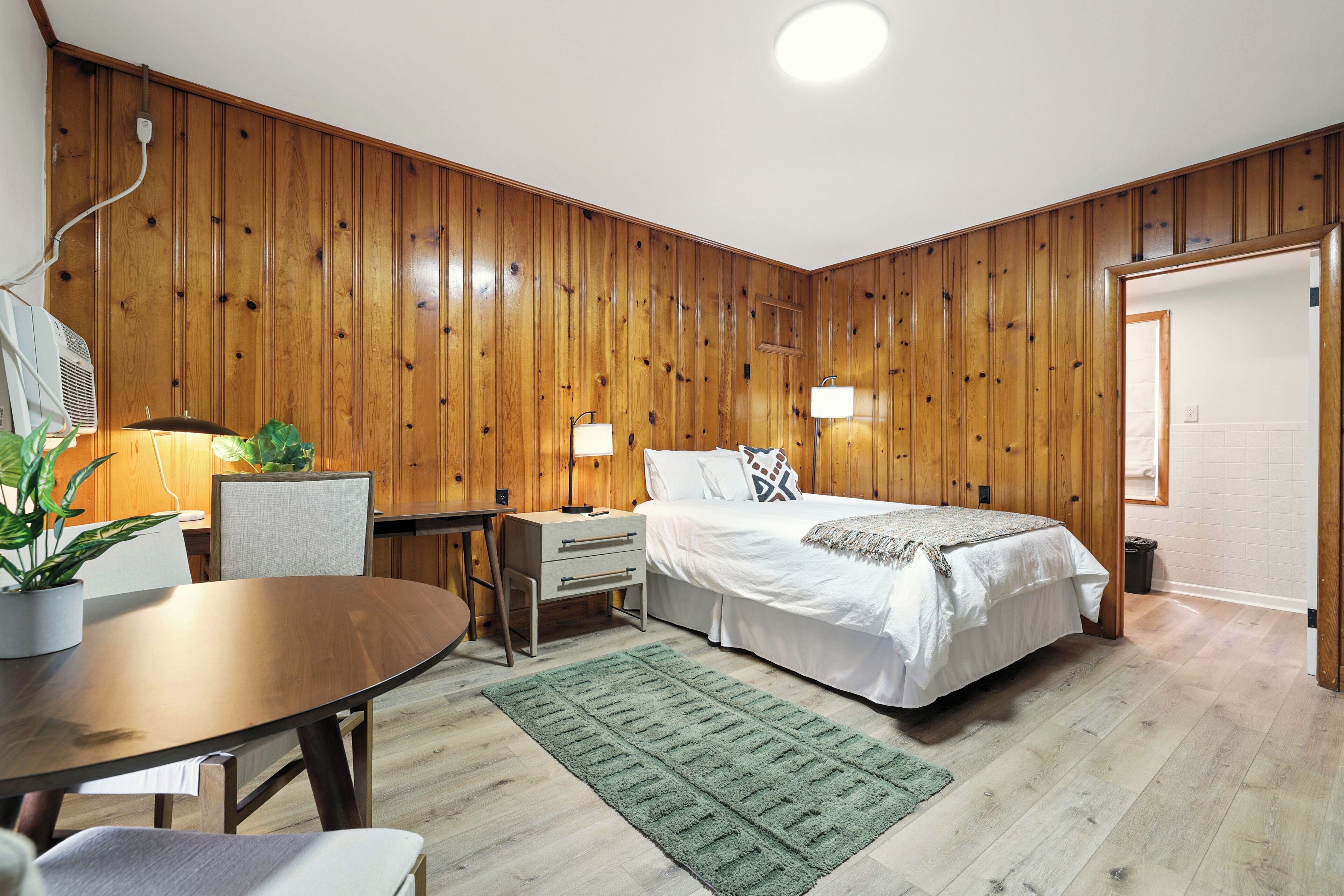 A room with wooden panel walls, a bed with white bedding, a desk with a chair, and a green rug.