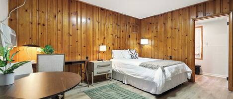 A room with wooden panel walls, a bed with white bedding, a desk with a chair, and a green rug.
