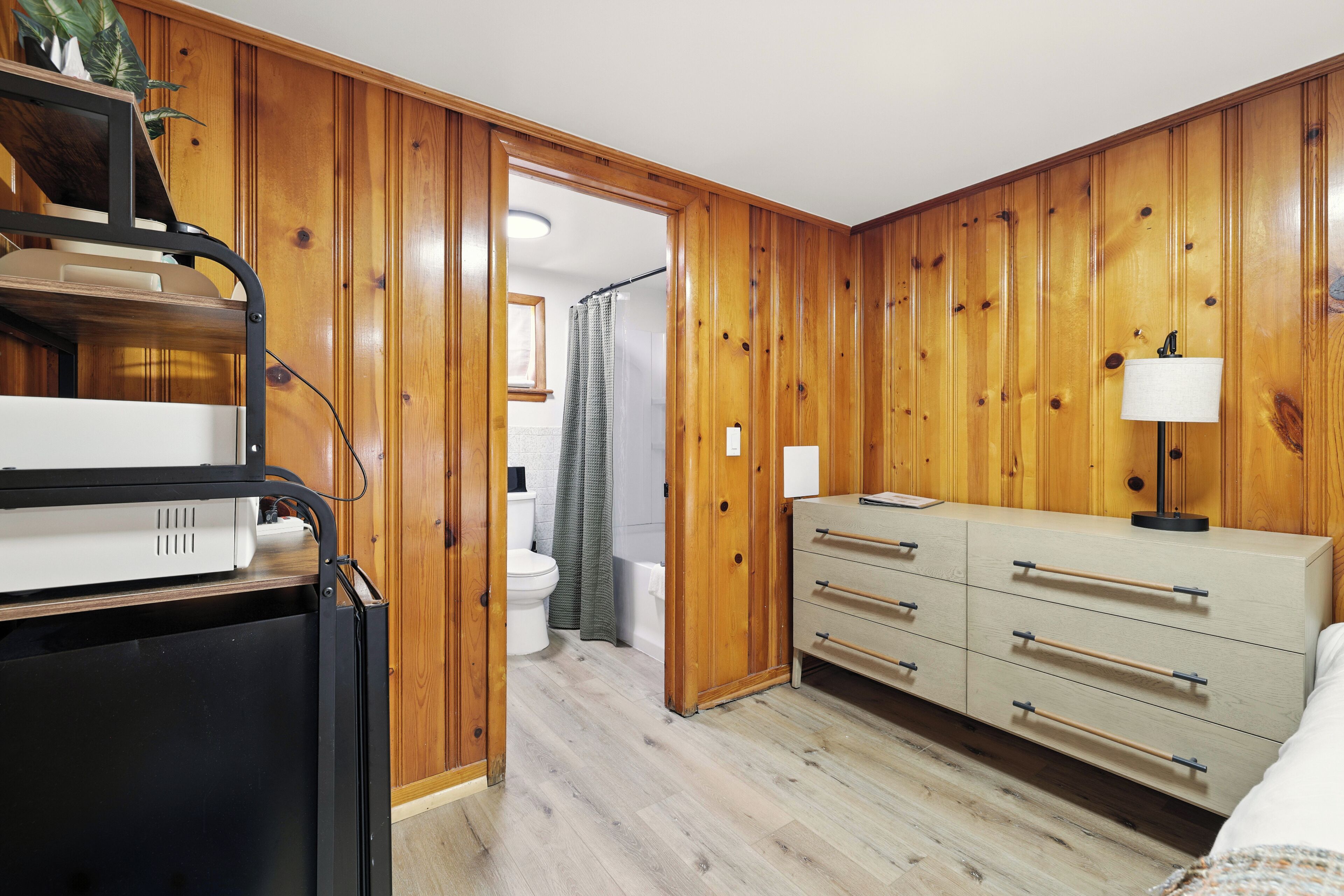 Ein Zimmer mit Holzwänden, einem Bett mit weißem Kopfteil, einem Schminktisch mit Schubladen, einer Lampe und einem Schreibtisch mit Computer und Pflanze.