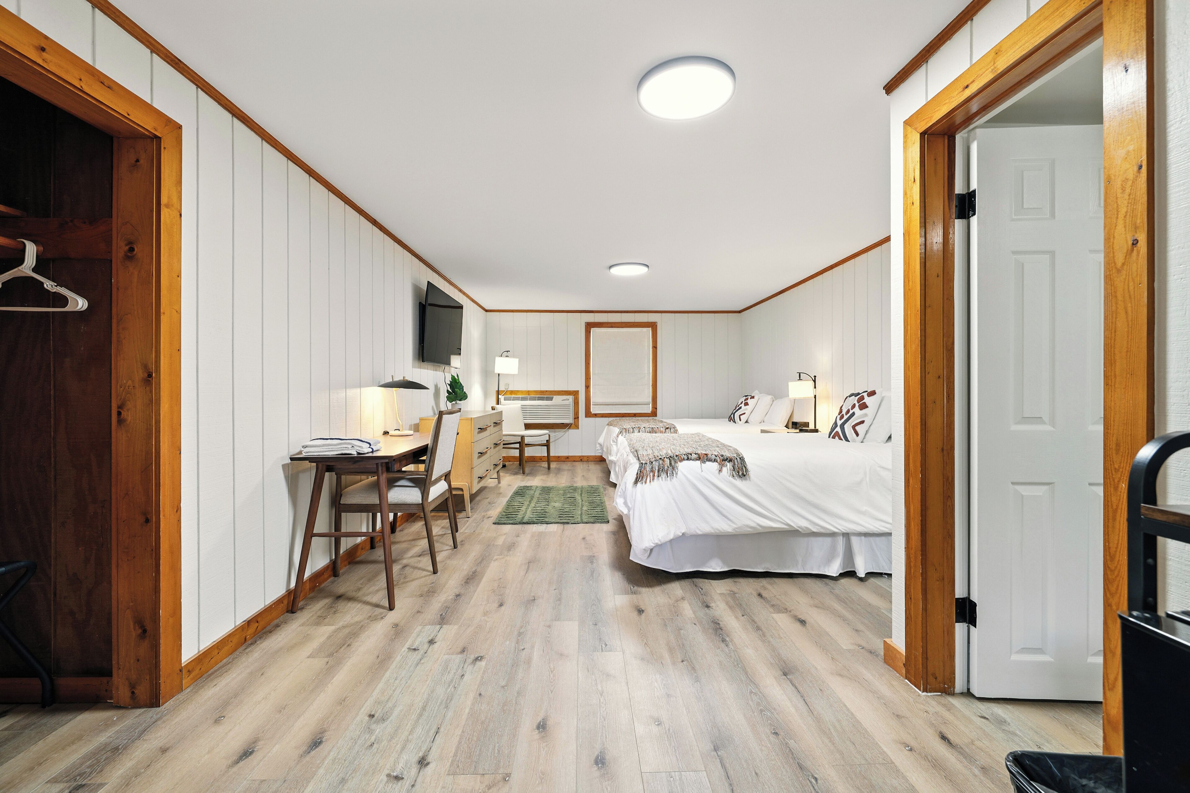 Un dormitorio con suelo de madera, un escritorio con silla, una cama con edredón blanco y un televisor montado en la pared.