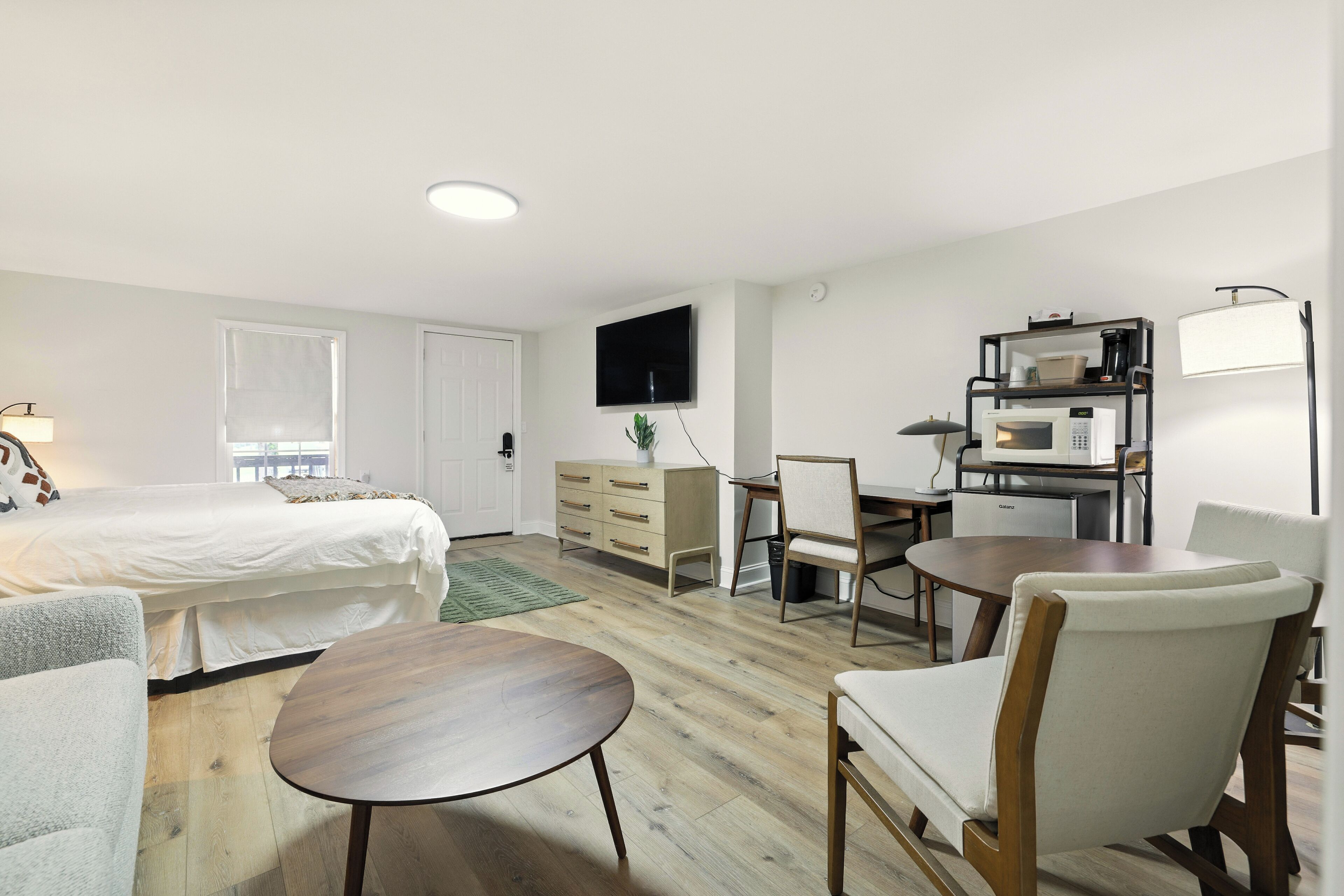 A hotel room with a bed, a desk, a chair, a table, a television, and a microwave.
