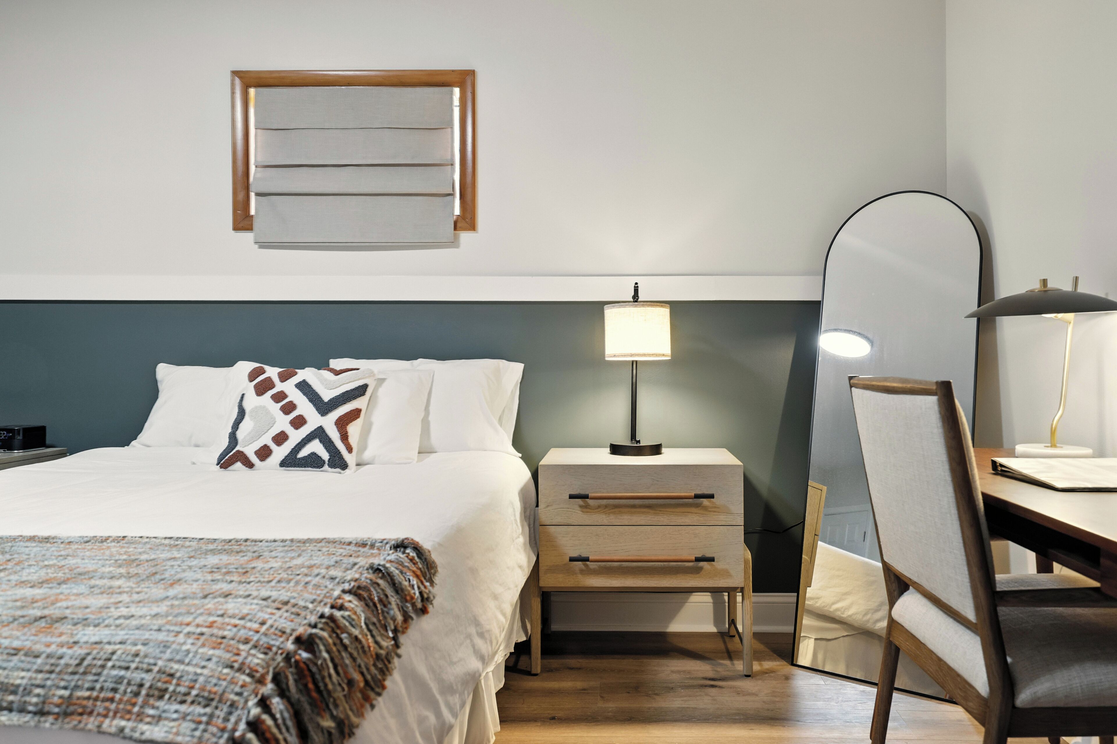 A neatly arranged hotel room with a bed, bedside table, lamp, chair, and a mirror.